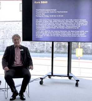 Bild: Thomas Abel im Anzug sitzt neben einem Screen bei einem Vortrag.