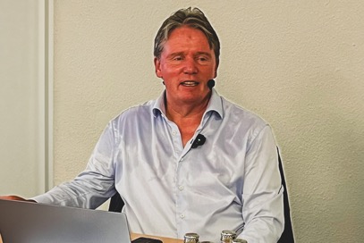 Bild: Thomas Abel sitzt hinter einem Tisch und hält eine Vorlesung mit einer Präsentation